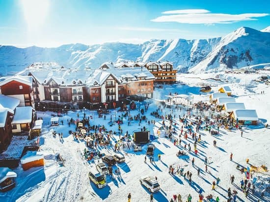 Aerial view of Gudauri ski resort village, snow-covered slopes, busy crowd, and Caucasus mountains under bright winter sun.