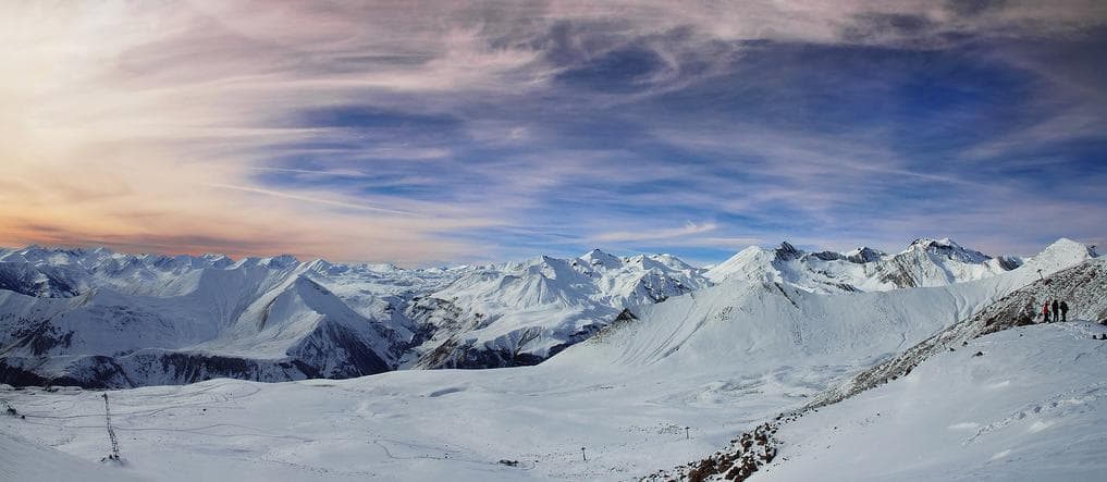 Panoramic winter view from Gudauri ski resort showing snow-covered Caucasus peaks and a colourful sunset sky, with a few skiers on a ridge.