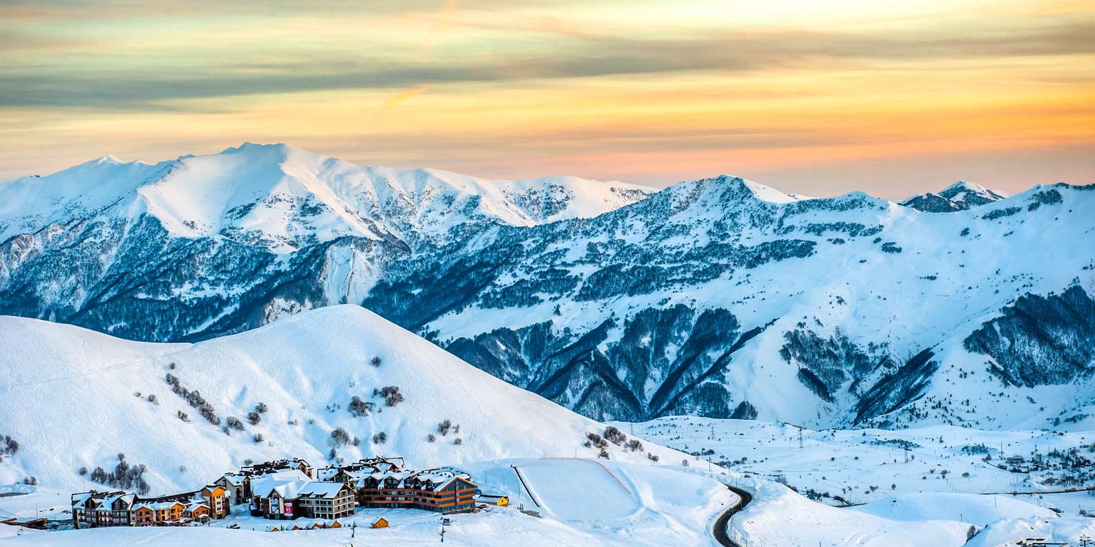 Gudauri ski resort village nestled in deep snow at sunset, with sweeping Caucasus mountain peaks glowing amber behind it